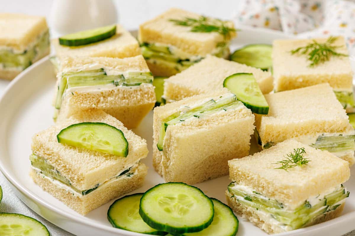 A plate of square cucumber tea sandwiches on white bread, garnished with fresh dill and cucumber slices, with extra cucumber slices and dill on the side.