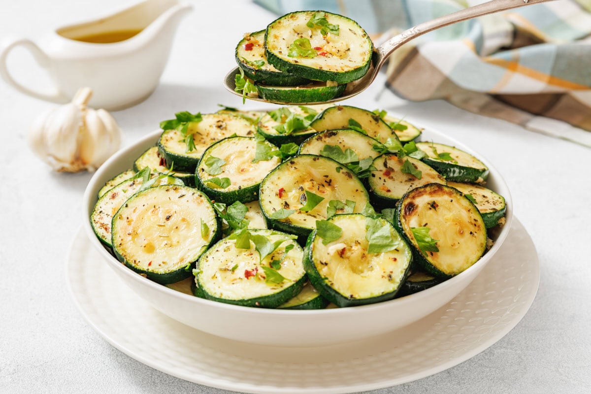 A white bowl filled with cooked, sliced zucchini garnished with herbs. A spoon holds several zucchini slices above the bowl. A garlic bulb and sauce boat are in the background.