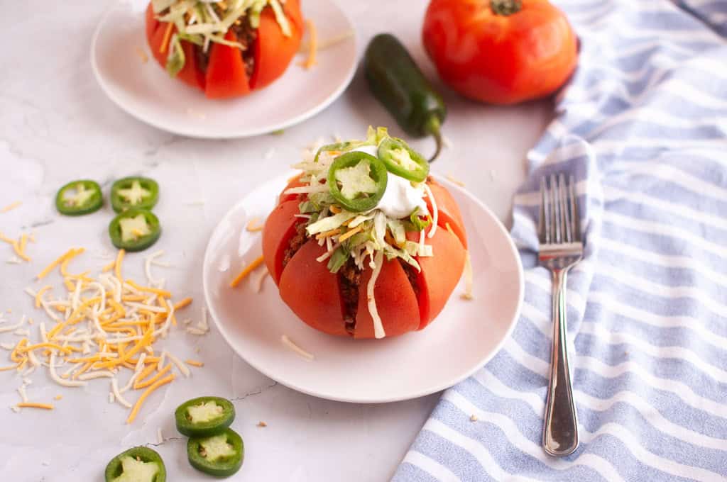 Two taco stuffed tomatoes on white plates with toppings and a fork.