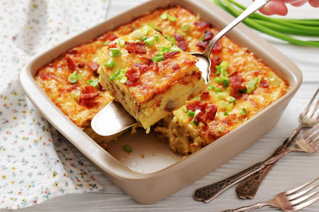 A baked casserole topped with chopped green onions and bacon is being served from a square dish with a metal spatula. Three forks lie beside the dish.
