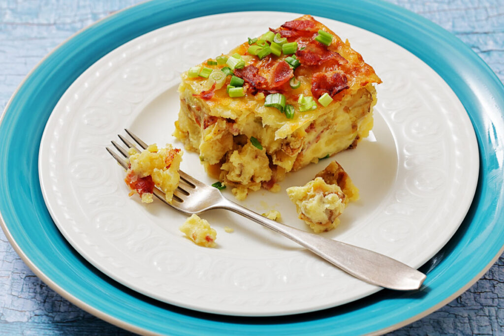 A square slice of twice-bake potato with chopped green onions and bacon sits on a white plate with a fork and a blue rim.