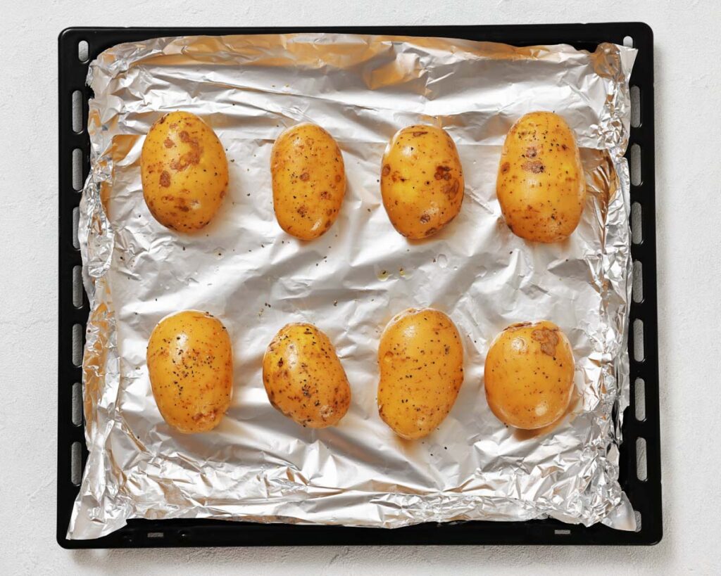 Eight whole, seasoned potatoes are arranged on a foil-lined baking tray, ready to be roasted in the oven.