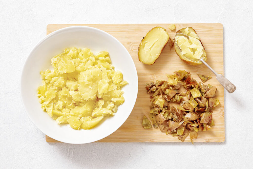 A bowl of mashed potatoes next to chopped potato peels and a spoon on a wooden cutting board.