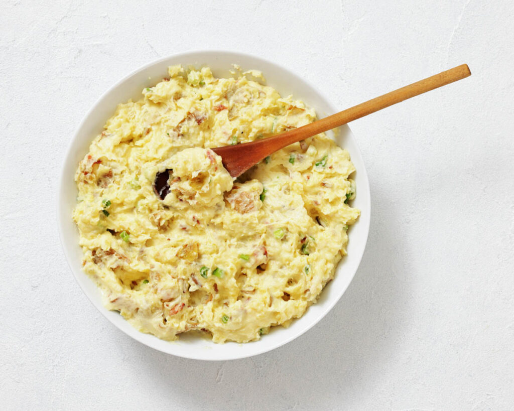 A white bowl filled with creamy potato salad, garnished with chopped herbs, with a wooden spoon resting inside.