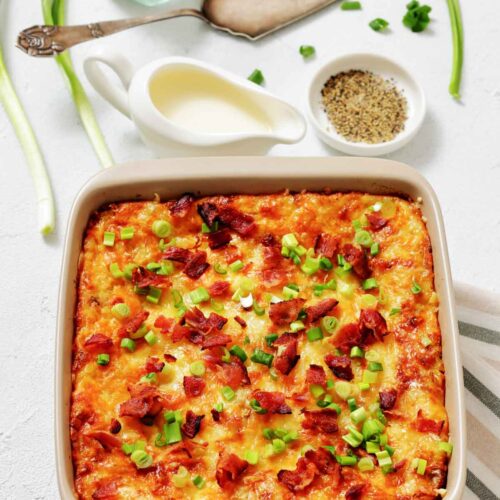 Baked casserole topped with chopped green onions and bacon in a square dish, with cream sauce, seasoning, and green onions beside it on a white surface.