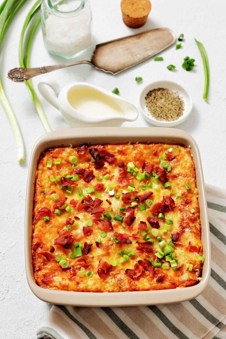Baked casserole topped with chopped green onions and bacon in a square dish, with cream sauce, seasoning, and green onions beside it on a white surface.