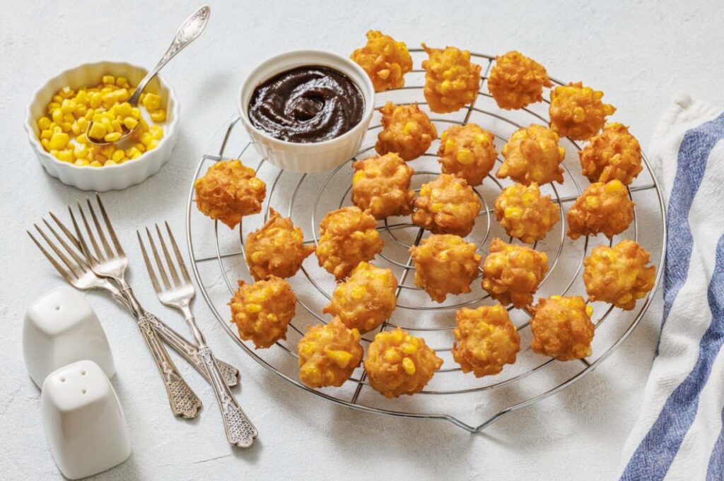 Golden corn fritters are arranged on a cooling rack with a small bowl of dipping sauce, a dish of corn, several forks, and salt and pepper shakers nearby.