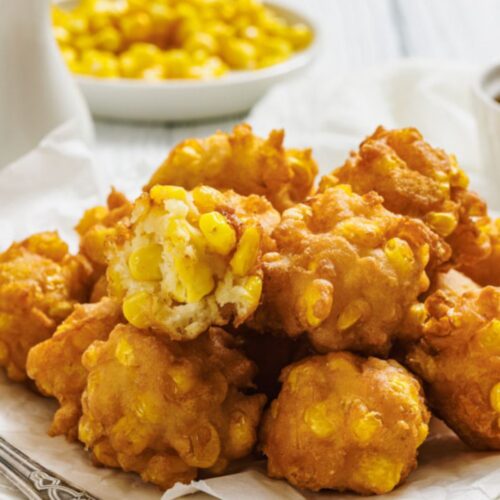 A plate of golden brown corn fritters sits on white parchment paper, with one fritter broken open to show the corn kernels inside; a bowl of corn is in the background.