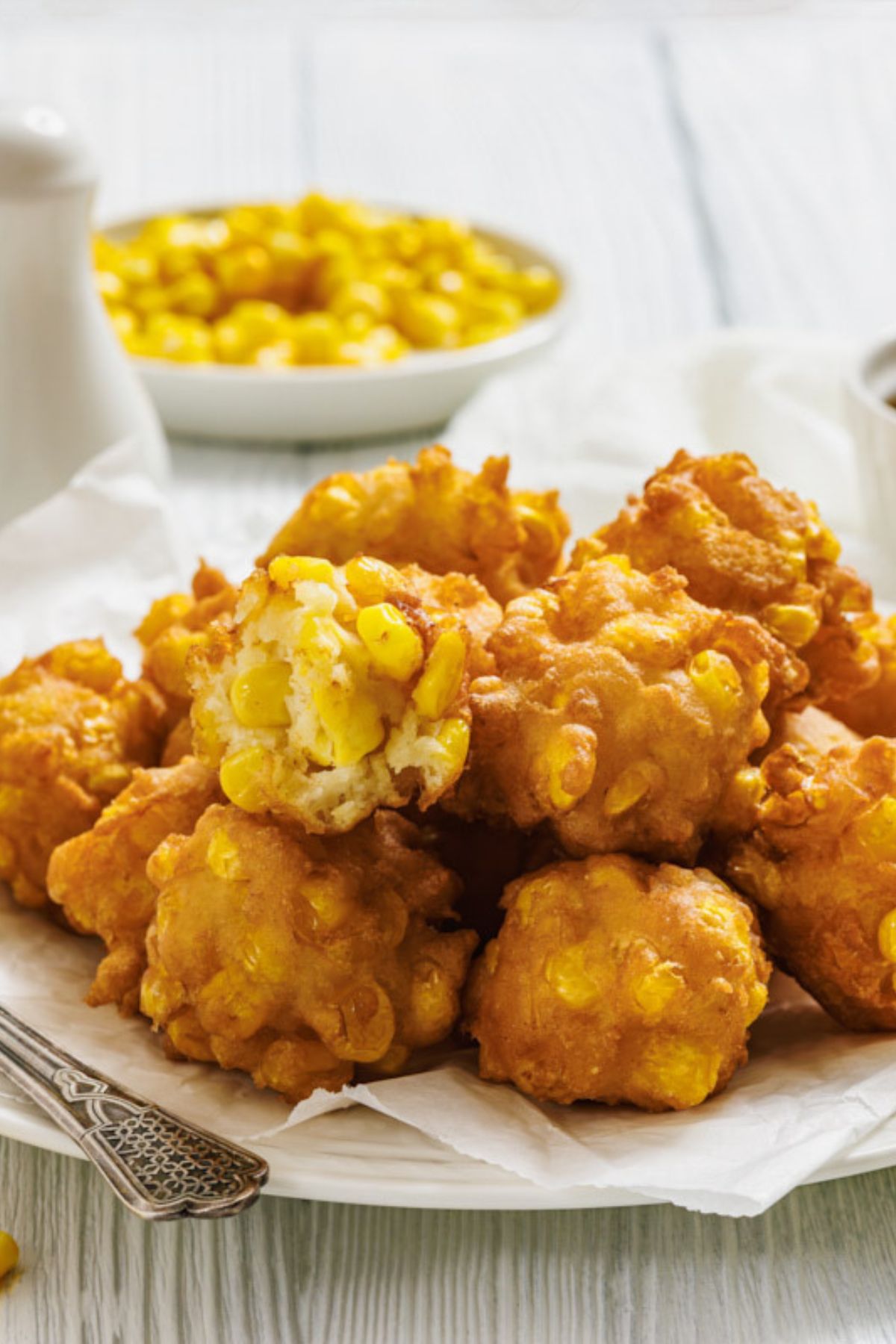 A plate of golden brown corn fritters sits on white parchment paper, with one fritter broken open to show the corn kernels inside; a bowl of corn is in the background.
