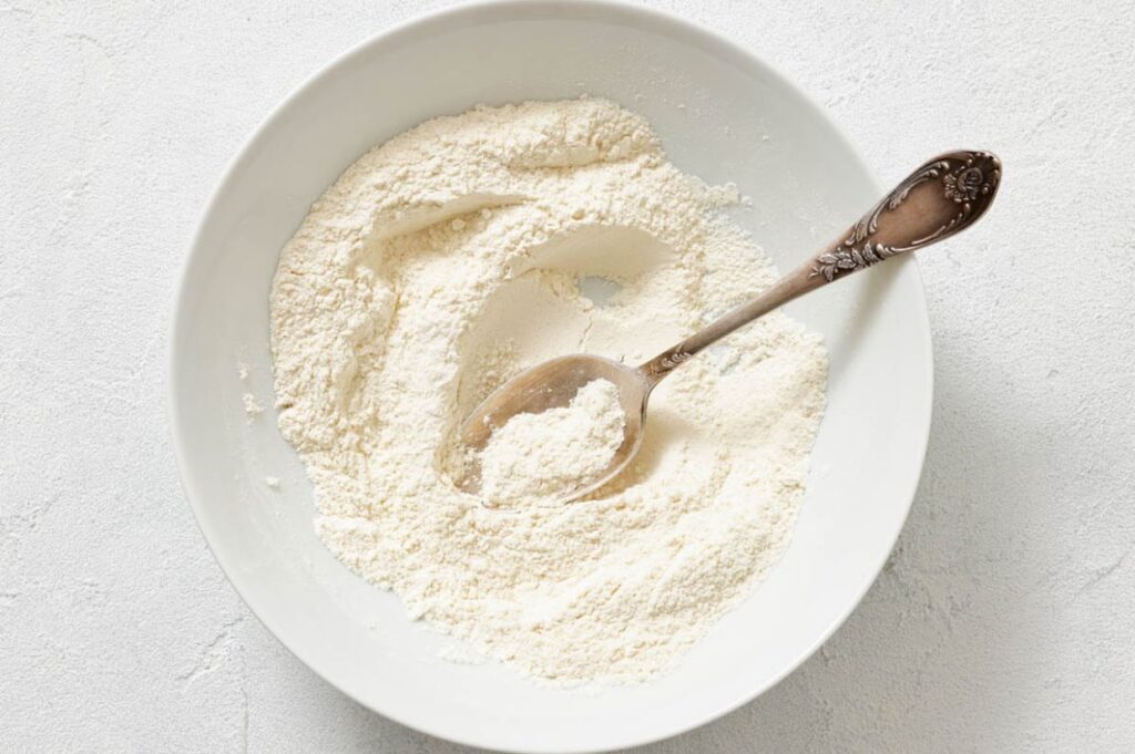 A white bowl filled with flour and a silver spoon resting inside, placed on a light-colored surface.