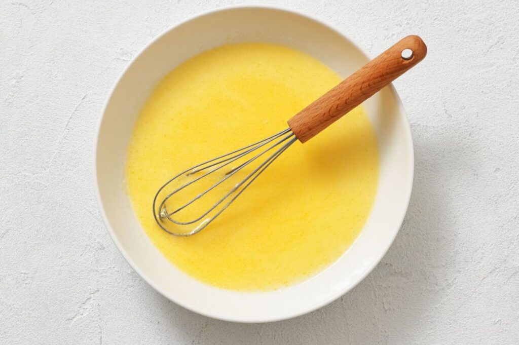 A metal whisk with a wooden handle rests in a white bowl containing a yellow liquid mixture on a light-colored surface.