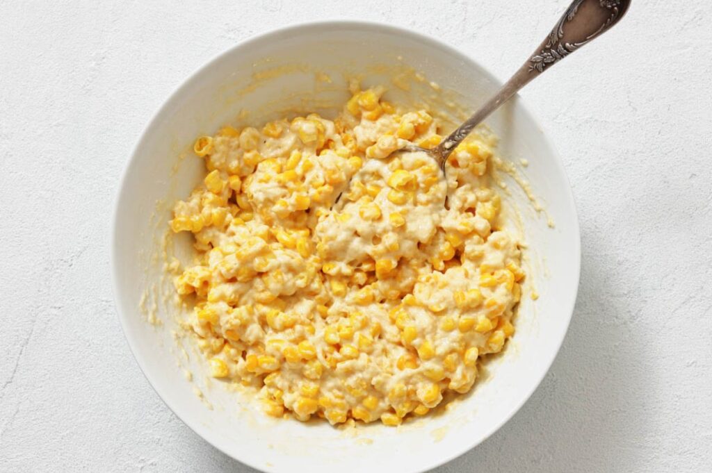 A white bowl filled with a creamy corn mixture, stirred with a silver spoon, sits on a light-colored surface.