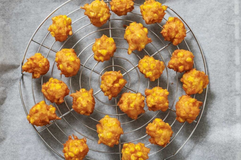 Several golden corn fritters are arranged on a round cooling rack set over parchment paper.