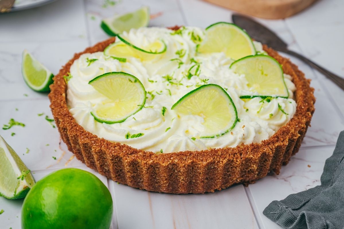 A key lime pie with a graham cracker crust, topped with whipped cream and lime slices, sits on a marble surface with lime wedges nearby.
