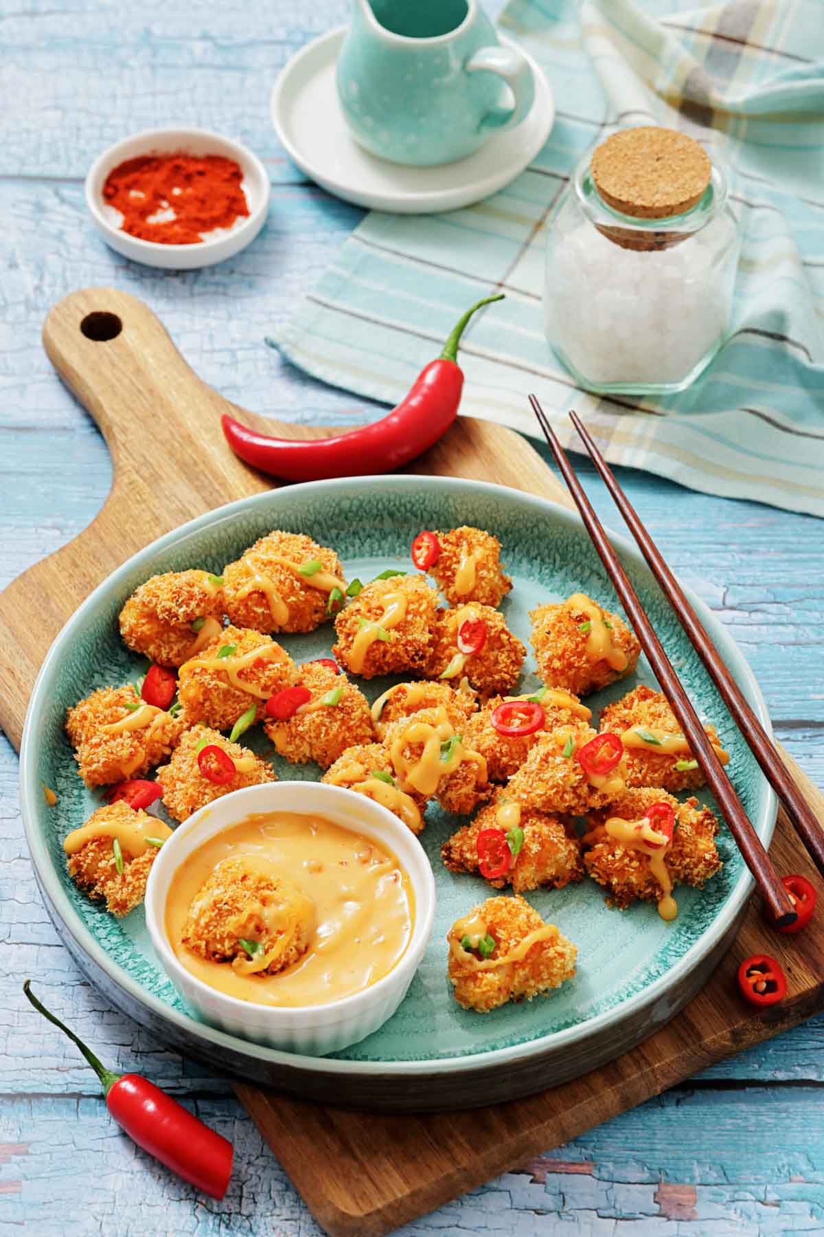 A plate of crispy breaded chicken bites garnished with sliced red chili and sauce, served with a dipping sauce; chopsticks and spices are arranged nearby on a blue wooden table.