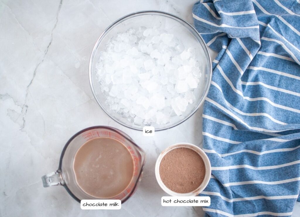 Overhead shot of a white marble surface with a bowl of ice, a measuring cup of chocolate milk, and a small bowl of hot chocolate mix, all labeled, with a blue and white striped napkin to the side.