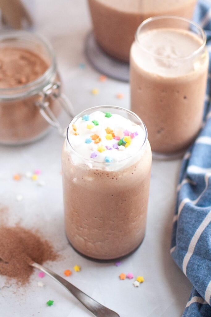 Two glasses of frothy brown frozen hot chocolate, one topped with whipped cream and colorful star sprinkles, on a white surface with scattered sprinkles and a jar of chocolate mix.