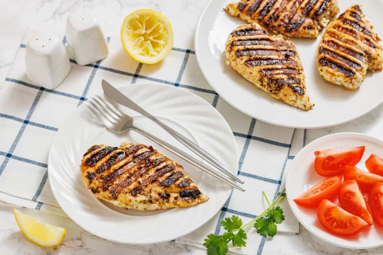 Grilled chicken breast on a white plate with fork and knife, sliced tomatoes, lemon halves, cilantro, and salt and pepper shakers on a checkered cloth.
