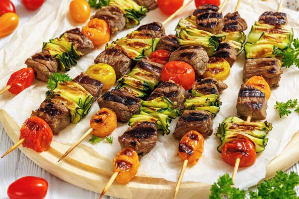 Grilled skewers with beef, cherry tomatoes, zucchini slices, and yellow tomatoes on a parchment-lined wooden board, garnished with parsley.