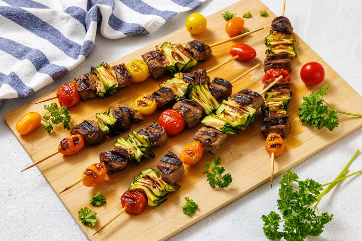 Grilled skewers with beef, zucchini, and cherry tomatoes on a wooden board, garnished with fresh parsley. A striped cloth and loose cherry tomatoes are nearby.