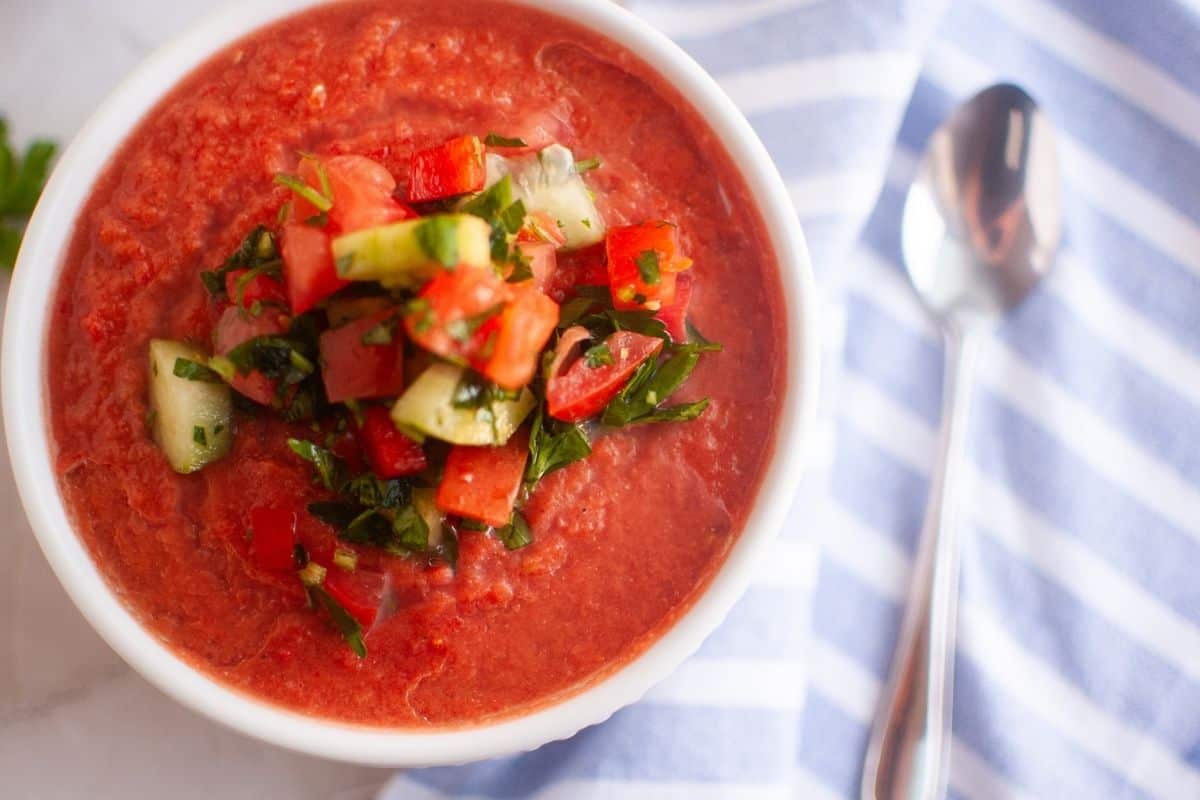 A bowl of classic gazpacho topped with diced cucumber, tomato, and herbs sits next to a striped cloth napkin and a spoon.