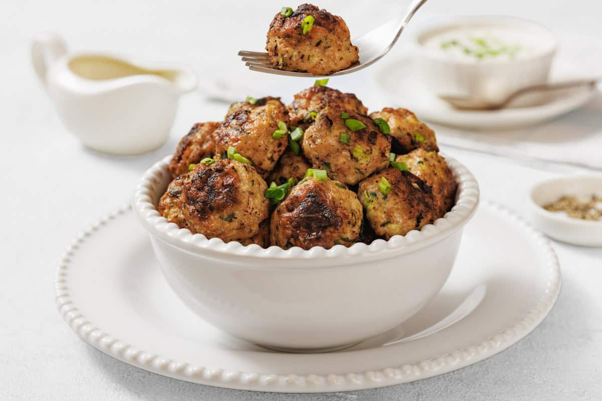 A white bowl filled with Chicken Zucchini Poppers garnished with chopped herbs, with a fork holding one meatball above the bowl.