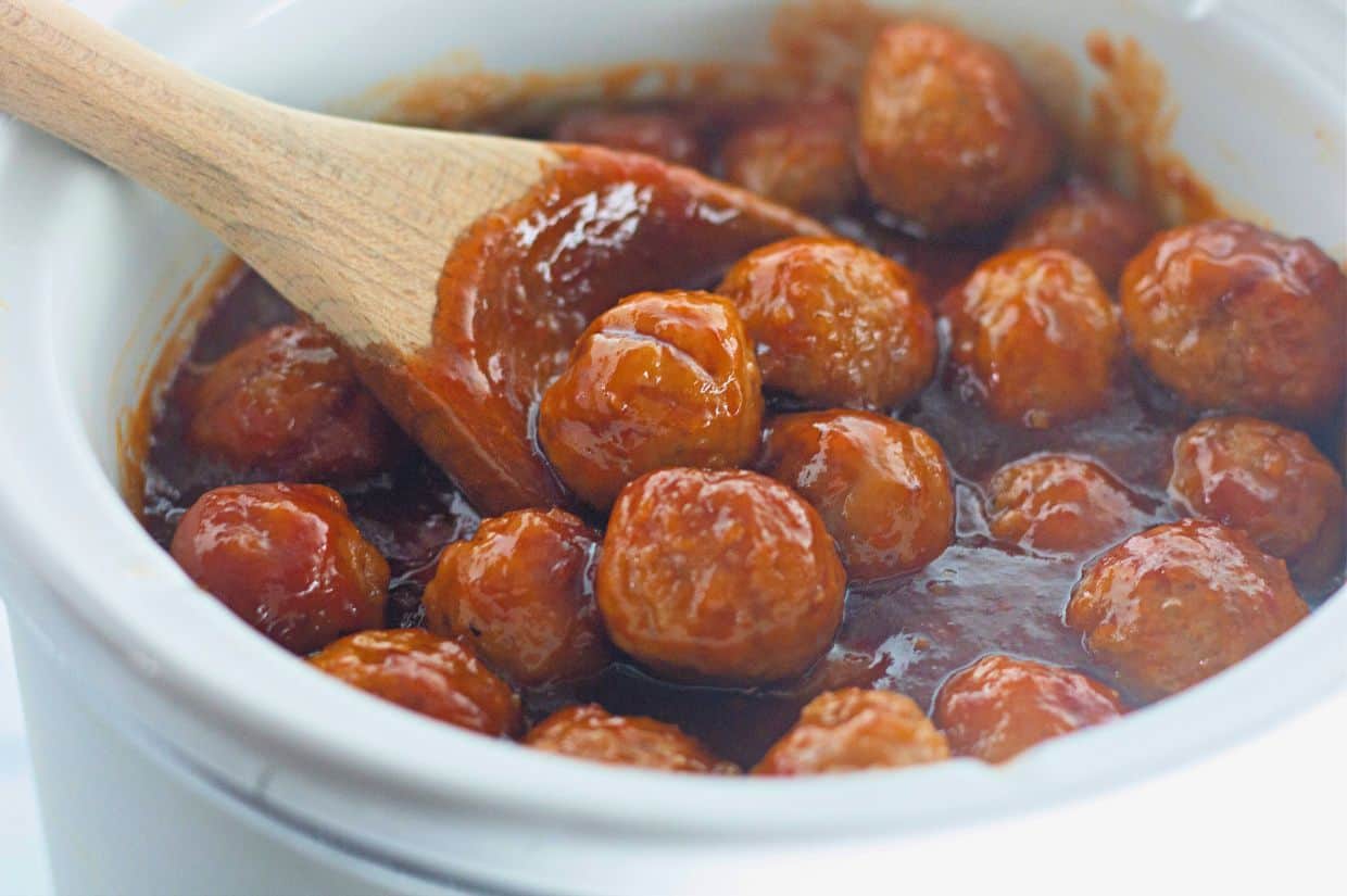 Honey-Garlic  Meatballs in a thick red sauce are being stirred with a wooden spoon in a white ceramic pot.