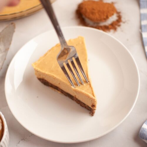 A hand holds a fork over a slice of layered dessert, possibly cheesecake, on a white plate with a spoonful of brown powder and striped napkin in the background.