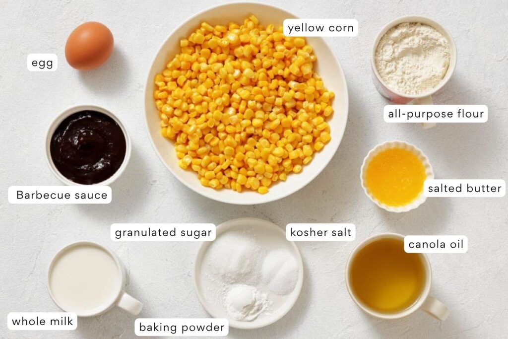 A variety of labeled ingredients on a white surface include an egg, yellow corn, flour, barbecue sauce, butter, sugar, salt, canola oil, milk, and baking powder.