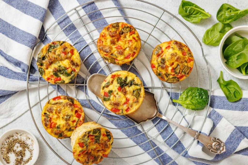 Six mini frittatas with vegetables on a round cooling rack, with spinach leaves, a bowl of pepper, and a striped cloth nearby.