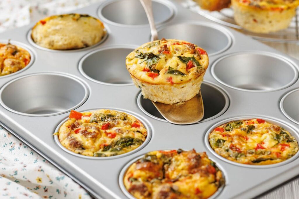 A muffin tin filled with baked egg muffins containing chopped vegetables, with one muffin being lifted out on a spatula.