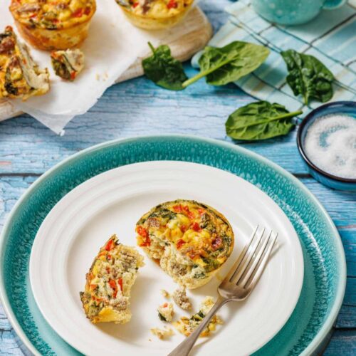 A plate with a vegetable egg muffin, partially eaten with a fork, sits on a blue table next to more egg muffins, spinach leaves, and a small jug.