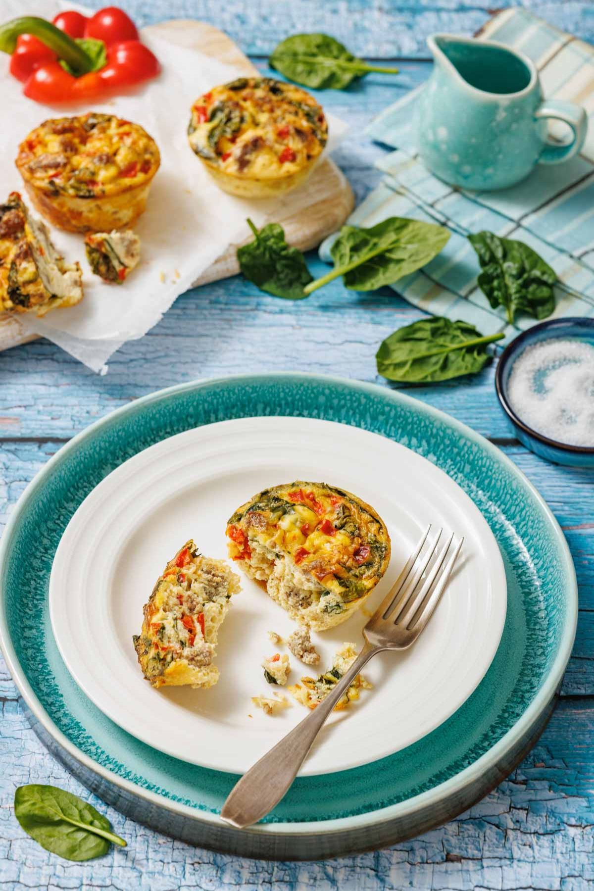 A plate with a vegetable egg muffin, partially eaten with a fork, sits on a blue table next to more egg muffins, spinach leaves, and a small jug.