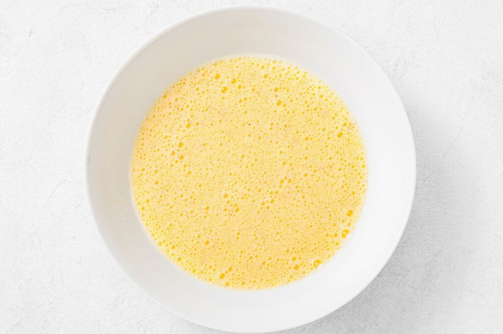 A white bowl filled with a frothy, yellow liquid mixture, likely beaten eggs, set on a light-colored surface.