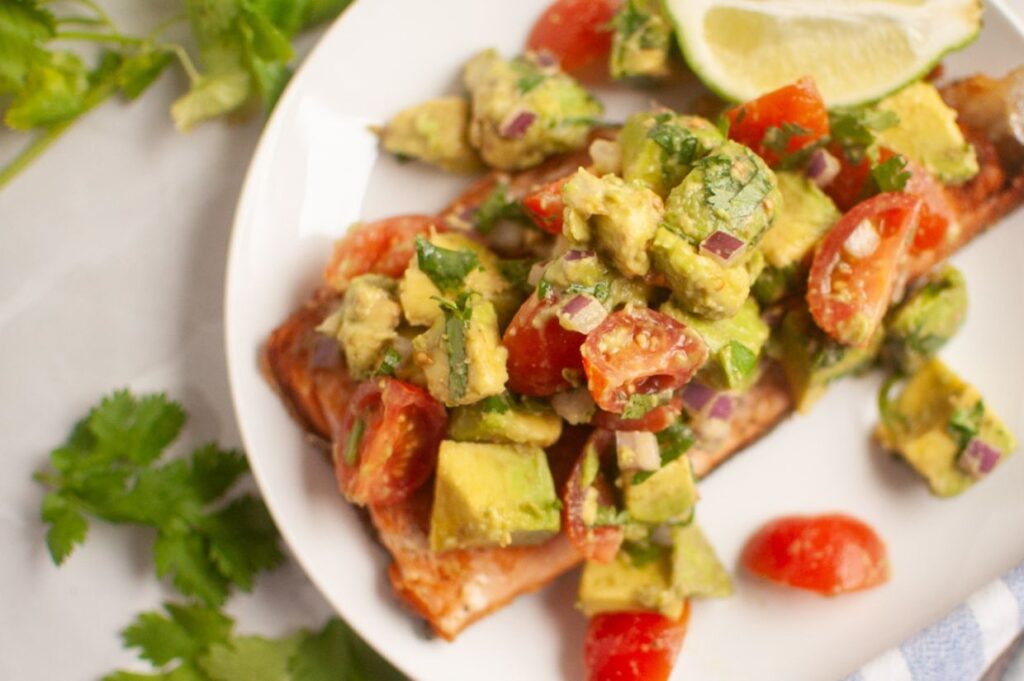 Grilled salmon fillet topped with avocado, tomato, red onion, and cilantro salsa on a white plate, garnished with a lime wedge.