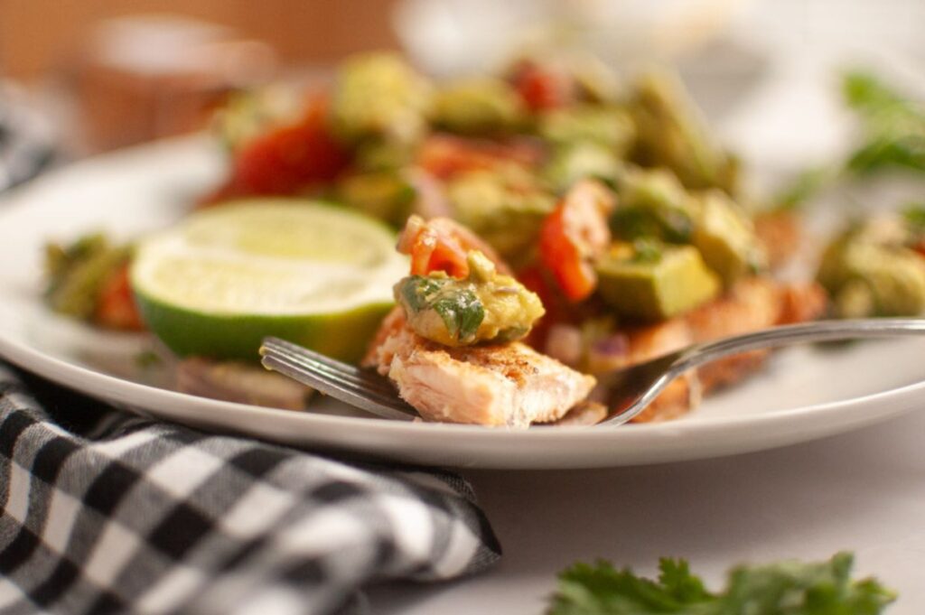 A close-up of a forkful of seared salmon topped with avocado and tomato, with lime slices on a white plate and a black-and-white checkered napkin nearby.