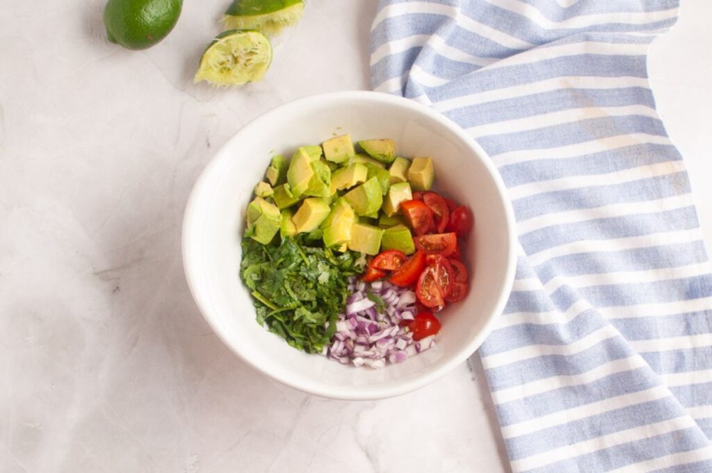 A white bowl with chopped avocado, cherry tomatoes, red onion, and cilantro on a marble surface, next to cut limes and a blue-striped cloth.
