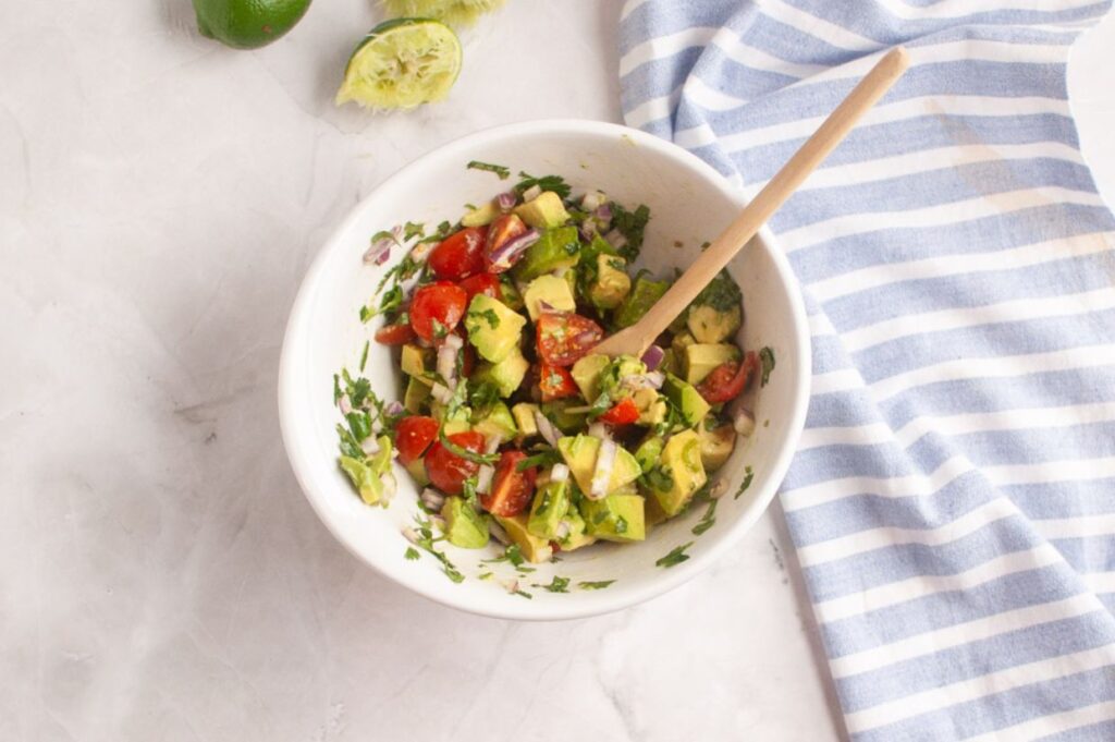 A white bowl with chopped avocado, tomato, red onion, and herbs, mixed with a wooden spoon, sits on a marble surface next to a blue striped towel and squeezed limes.