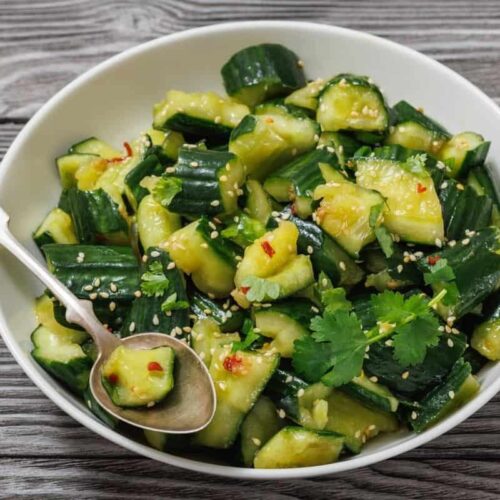 A white bowl filled with smashed cucumber salad garnished with cilantro, sesame seeds, and chili flakes, with a spoon resting inside. The bowl is on a wooden surface.