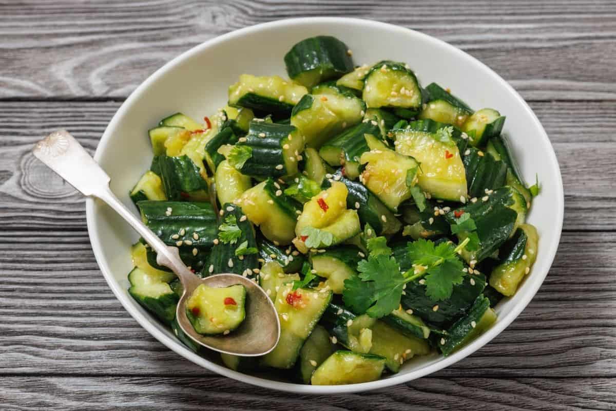 A bowl of smashed cucumber salad garnished with cilantro, sesame seeds, and chili flakes, with a spoon on the side, set on a wooden surface.