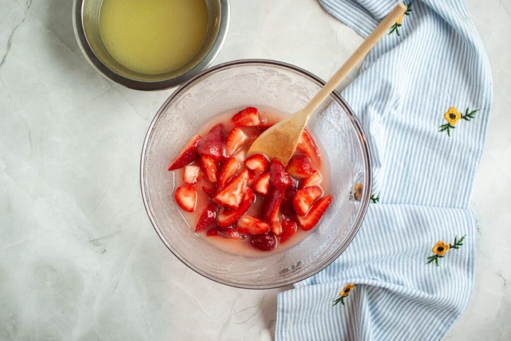 An overhead shot of a clear glass bowl containing sliced red strawberries partially submerged in a sugary liquid, with a wooden spoon resting in the bowl, a silver bowl of lemon juice to the upper left, and a blue and white striped napkin to the right.
