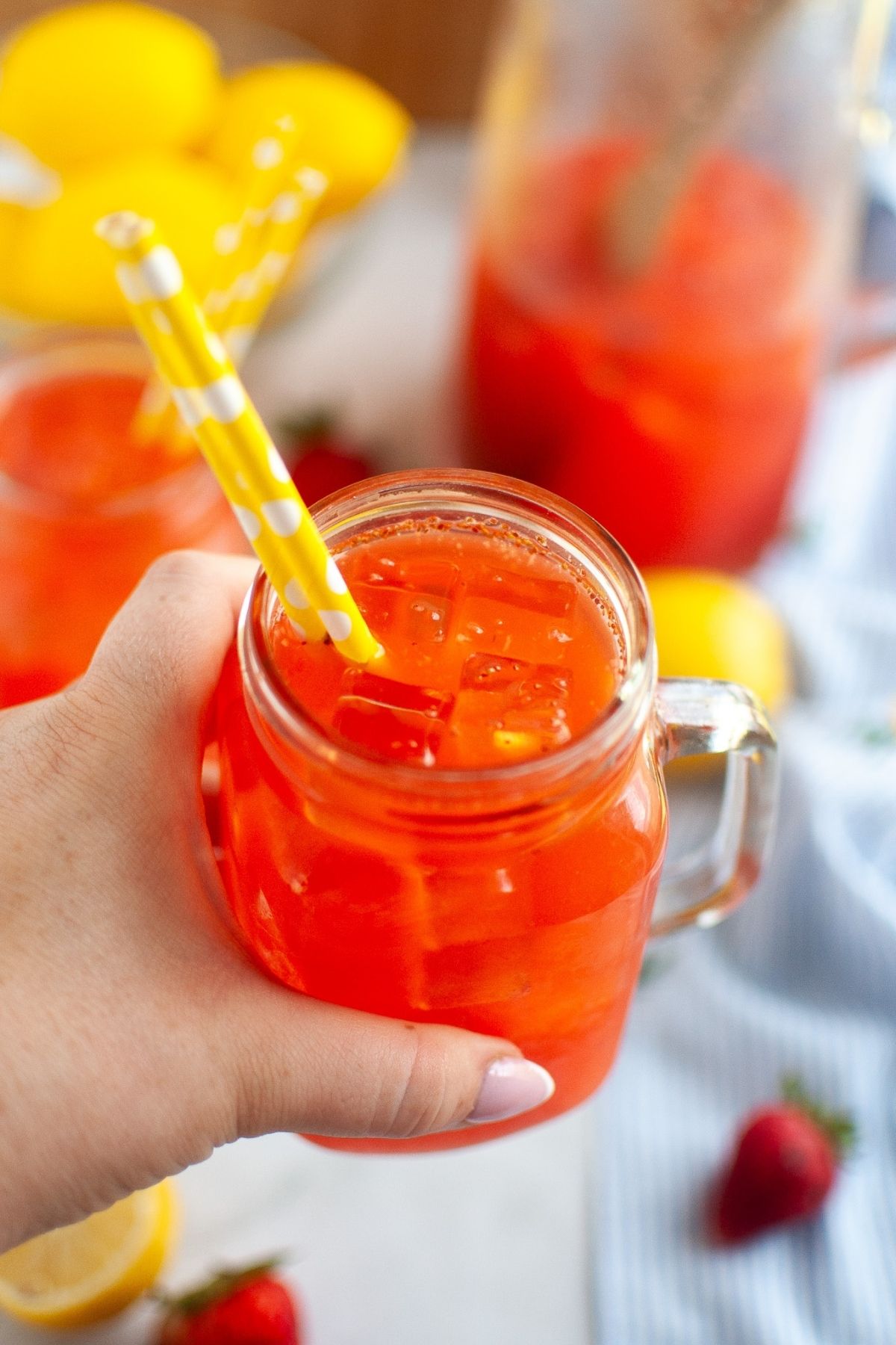 A hand holds up a clear mason jar filled with bright orange-red strawberry lemonade and ice cubes, with a yellow and white striped paper straw, against a blurred background of more lemons and a pitcher.