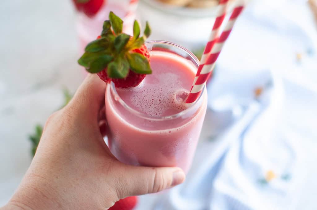 A hand holding a glass of pink strawberry milk with a red and white straw and a strawberry garnish.
