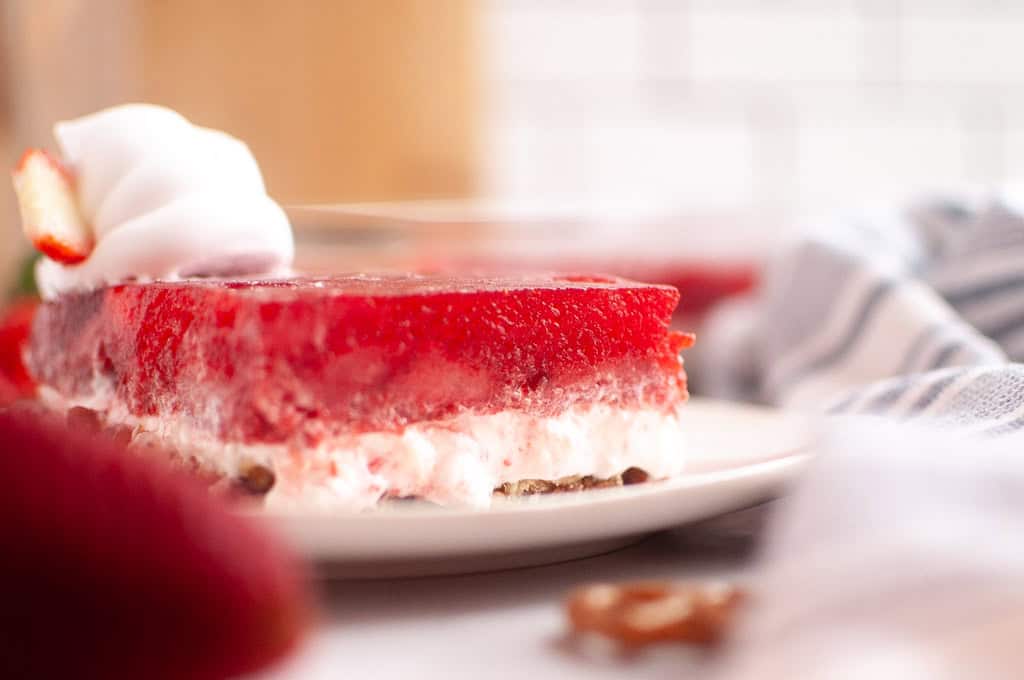 A slice of strawberry pretzel salad with whipped cream on a white plate, showing layers of red gelatin, creamy filling, and pretzel crust.