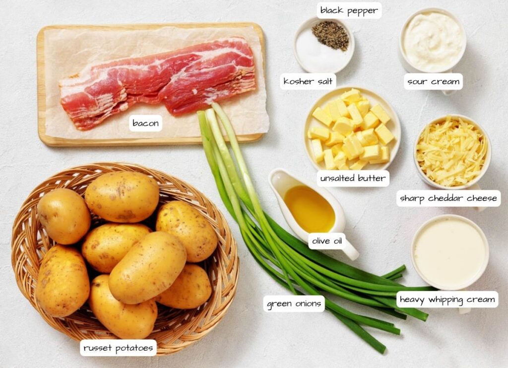 Ingredients for loaded mashed potatoes are arranged on a surface, including russet potatoes, bacon, green onions, olive oil, unsalted butter, cheddar cheese, sour cream, heavy cream, salt, and pepper.