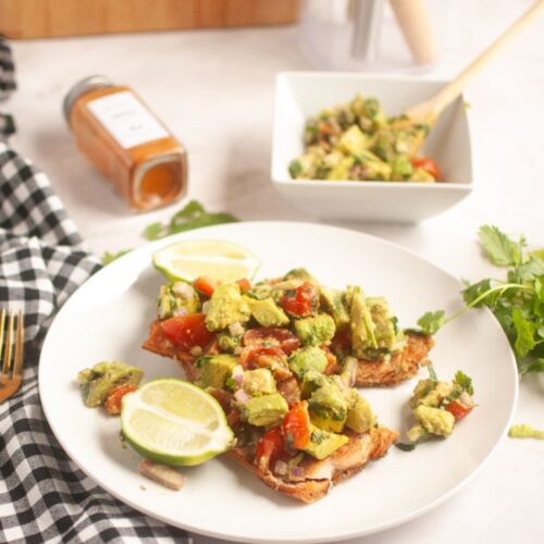 Plate with toasted bread topped with avocado and tomato mixture, garnished with lime slices. A bowl of the same mixture, a spice jar, and a checkered napkin are nearby.