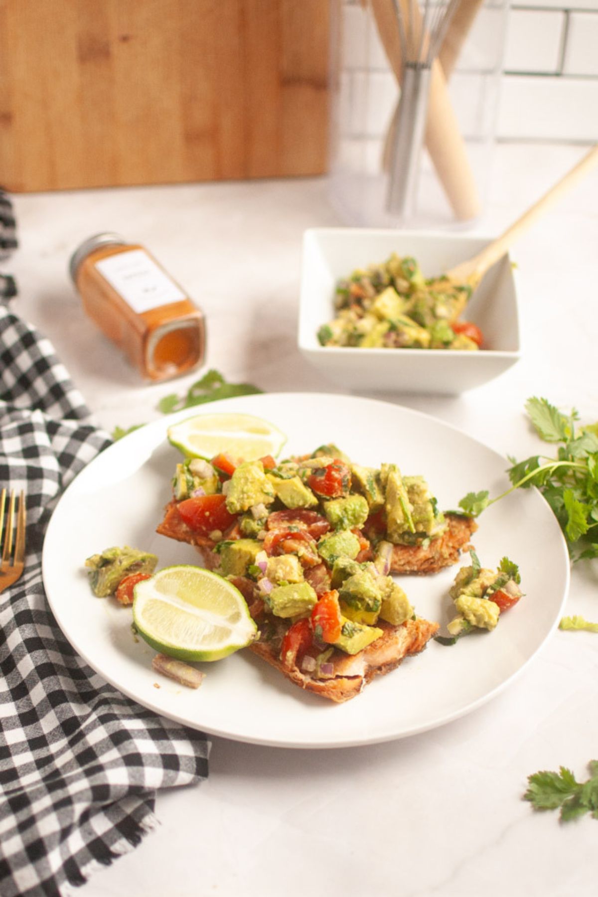 Plate with toasted bread topped with avocado and tomato mixture, garnished with lime slices. A bowl of the same mixture, a spice jar, and a checkered napkin are nearby.