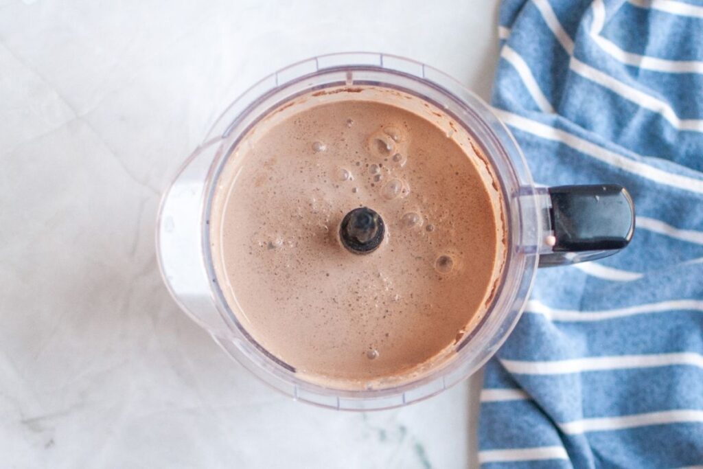 A top-down shot of a clear food processor bowl filled with a smooth, light brown, frothy liquid, which is blended frozen hot chocolate, on a white surface with a blue and white striped napkin.