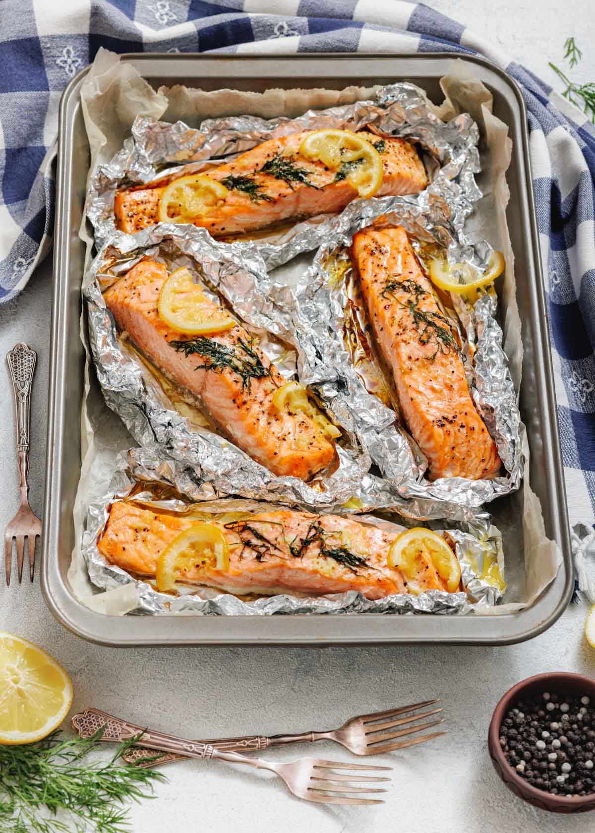 An overhead view of a metal baking pan lined with parchment paper, holding four individual foil packets of cooked salmon fillets, each garnished with lemon slices and fresh dill, with a blue and white plaid napkin to the left.