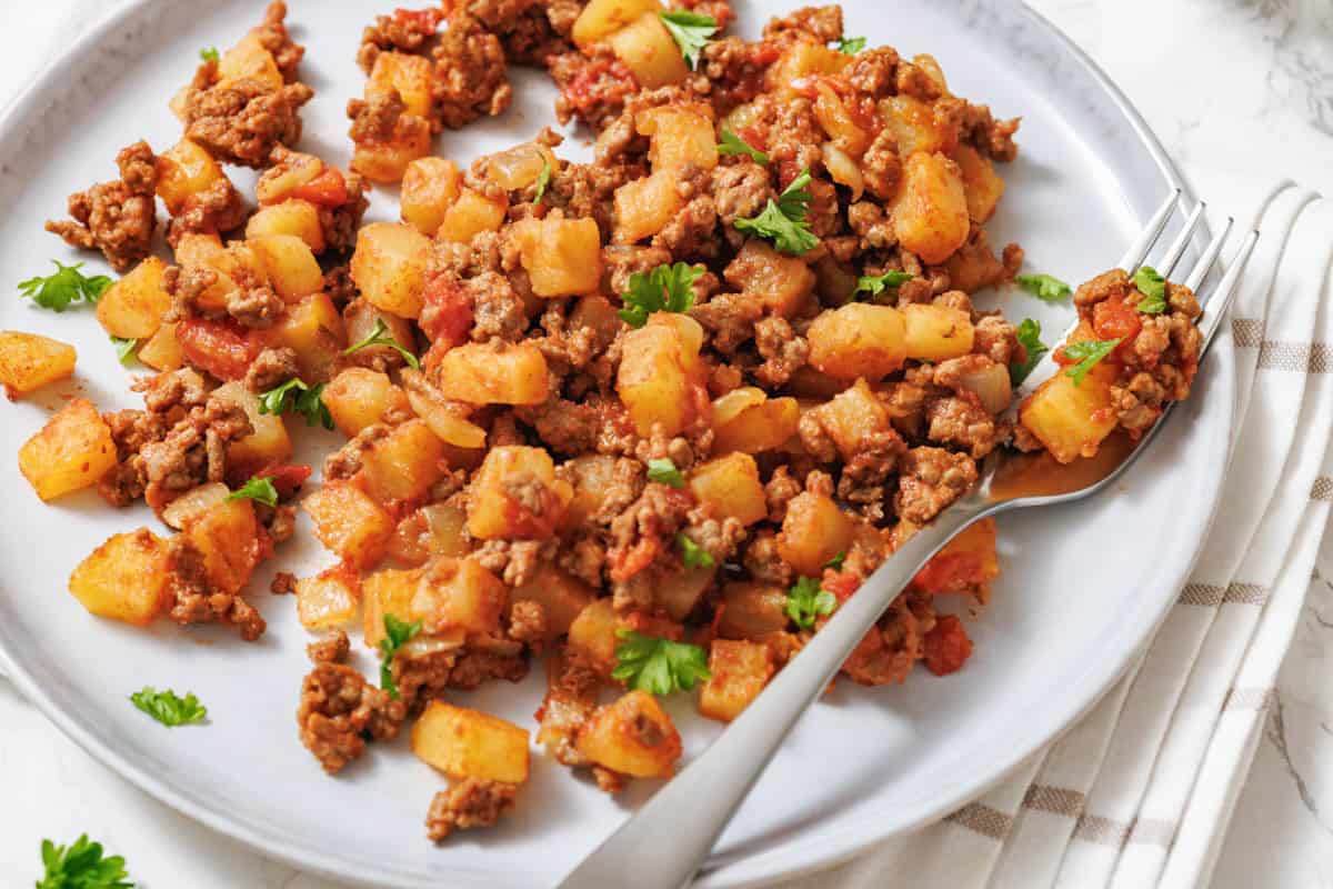 A plate of ground beef and diced potatoes cooked with tomatoes and herbs, garnished with parsley, with a fork on the side.